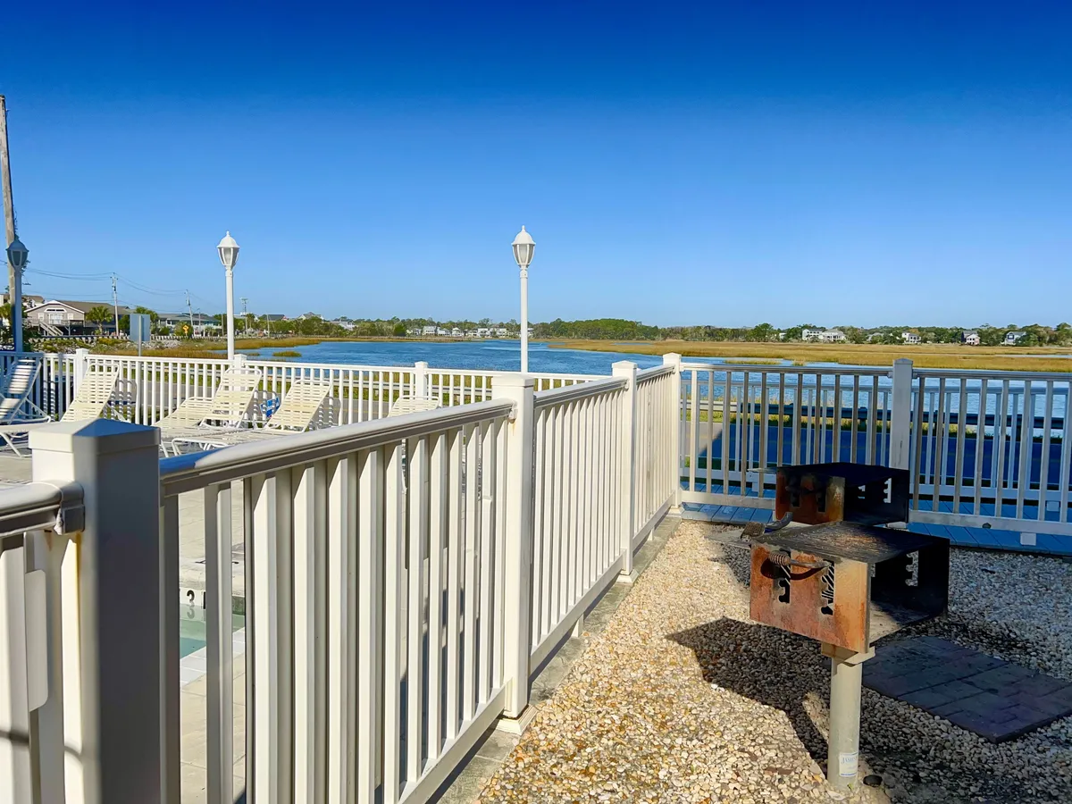 A waterfront patio scene complete with a grill, white railings, and lounge chairs, set against a scenic backdrop of the water. The area is ideal for barbecues, social gatherings, and relaxing afternoons by the sea.