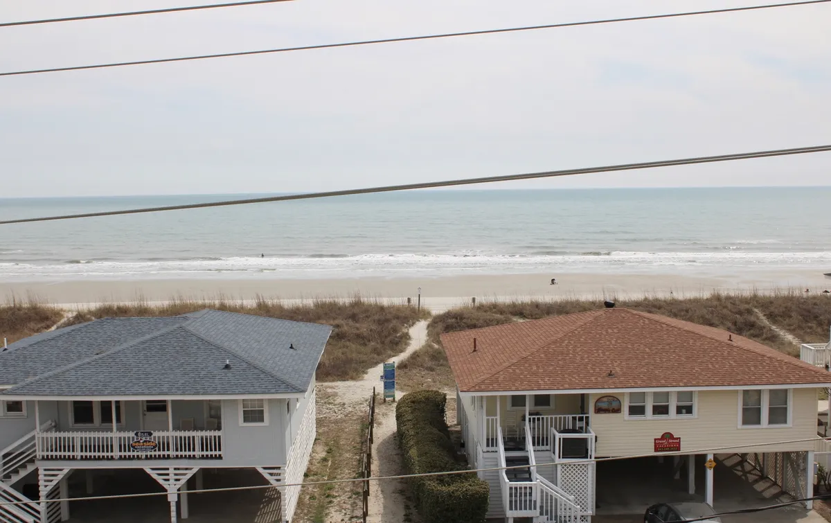 A peaceful view of two beach houses with a clear pathway leading directly to the sandy beach and the ocean beyond. The tranquil setting captures the essence of easy beach access, inviting sunrise walks and playful days building sandcastles mere steps from your door.