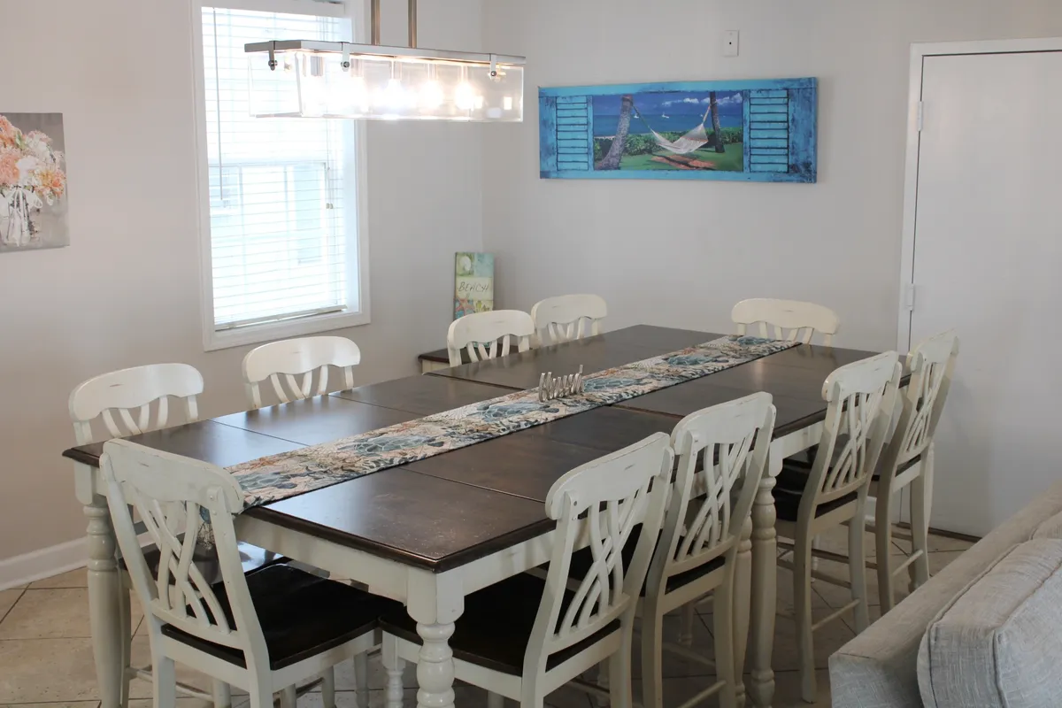 A long wooden dining table sits beneath a glowing chandelier, surrounded by decorative wall art and blue artwork. The table is perfectly set up for large family dinners, celebrations, or casual get-togethers.