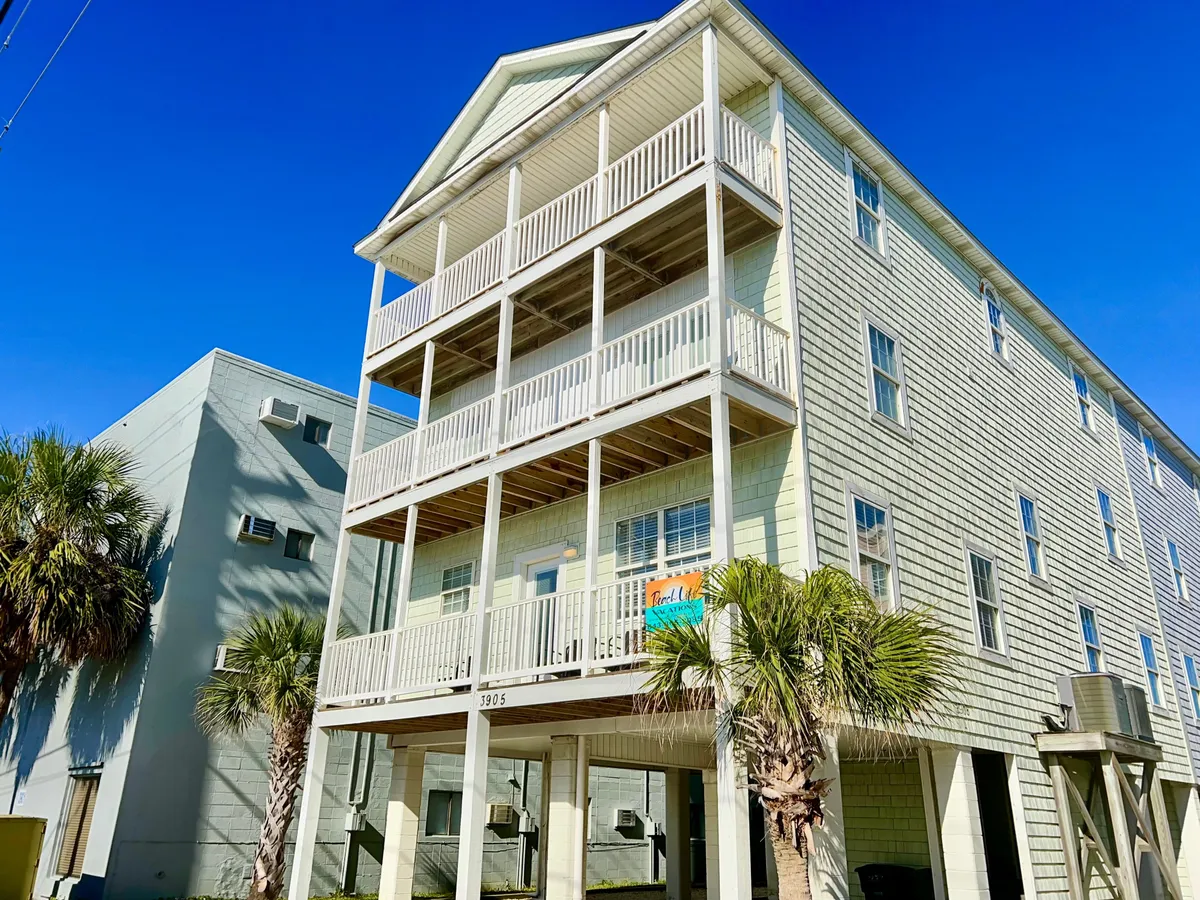 The image shows the exterior architecture of the property, framed by lush palm trees and featuring multiple balconies. The scene evokes a sense of arrival and togetherness, hinting at social moments enjoyed on the balconies and in the shared outdoor spaces. The cheerful mood highlights the vacation atmosphere and the property's proximity to beachside and local attractions.