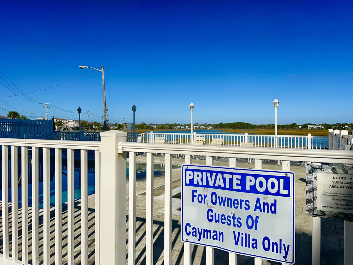 A private pool is showcased under a bright blue sky, bordered by a privacy fence and offering views of tranquil water beyond. The scene suggests a leisurely, exclusive retreat ideal for quiet mornings or social gatherings with friends.