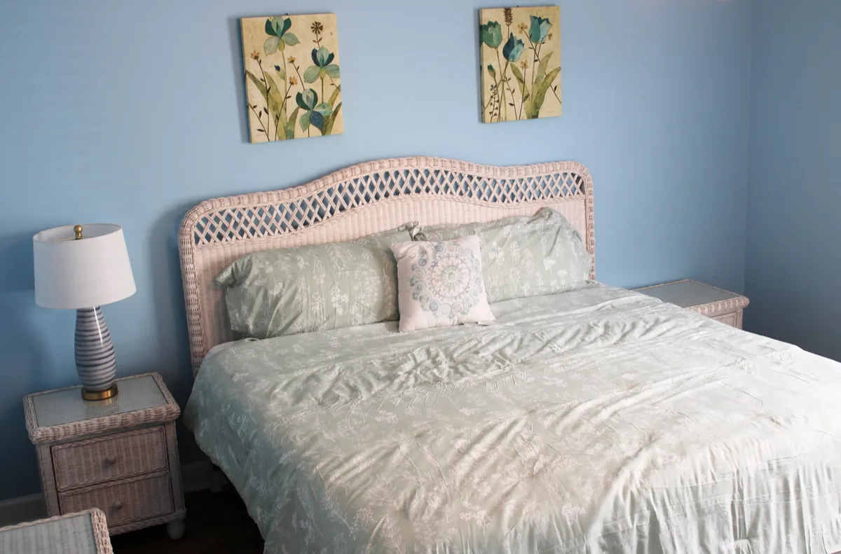 A serene bedroom featuring a bed with light green bedding, a wicker headboard, decorative pillows, lamps on bedside tables, and floral wall art. The space is softly lit and conveys a tranquil, welcoming atmosphere.