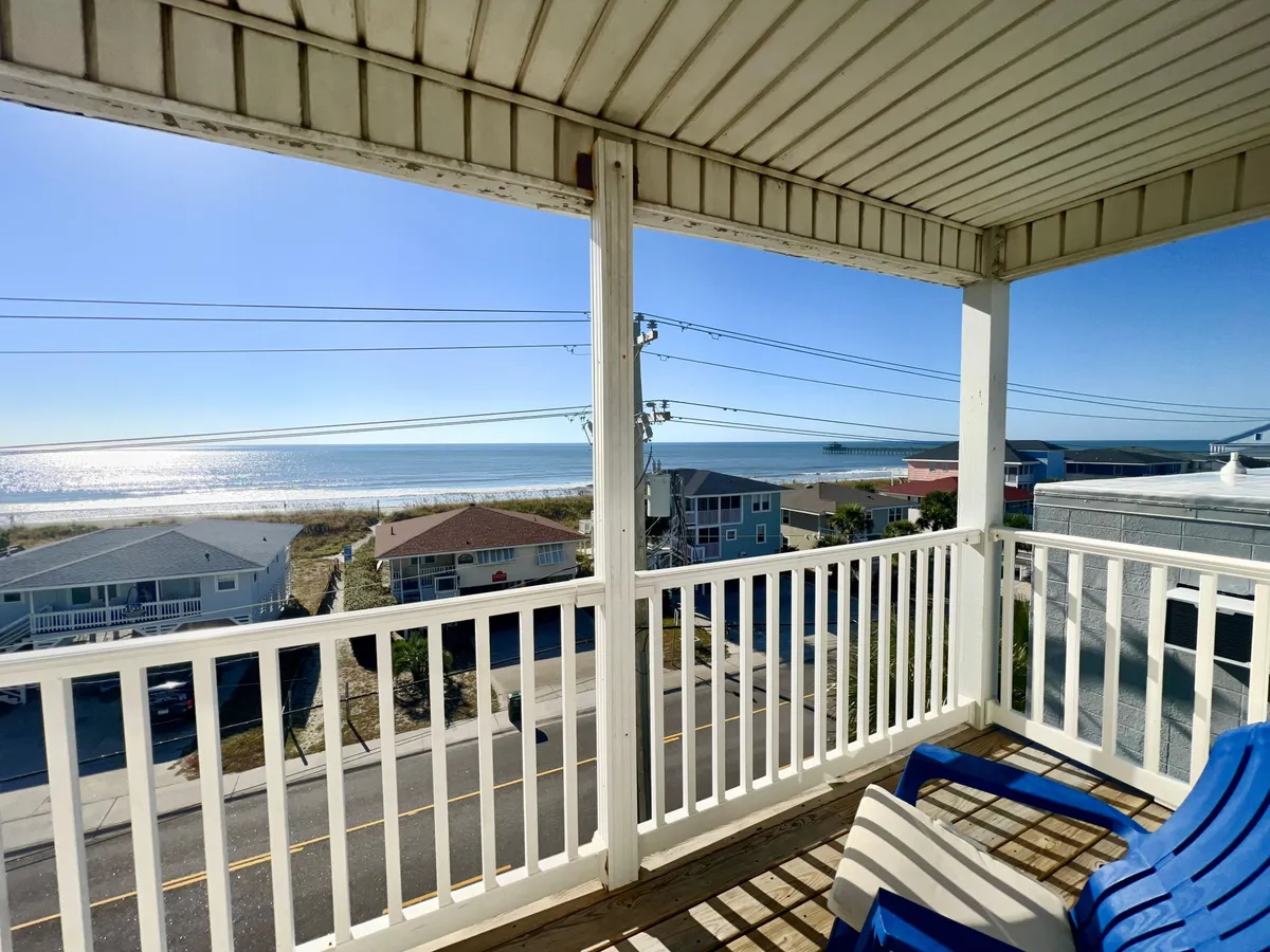 This image captures the view from a balcony overlooking the ocean, with a sturdy railing in the foreground and distant beach houses dotting the coastline. The tranquil scene shows a person relaxing on the balcony, immersed in the ocean breeze and the soft light of either sunrise or sunset. The expanse of water and sky emphasizes a seamless indoor-outdoor connection.