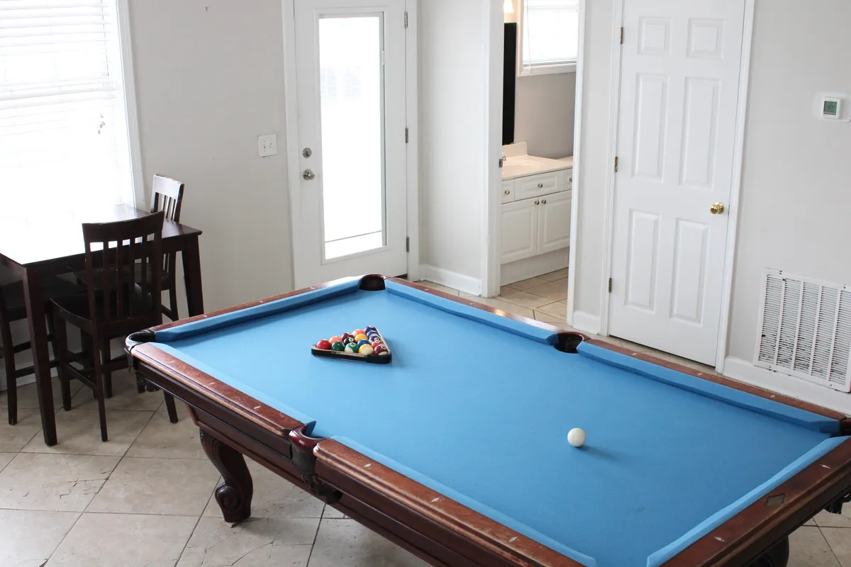 This image showcases a spacious interior area featuring a pool table, a dining table, and a visible doorway. The scene captures people enjoying a friendly game of pool and others gathered around the table, suggesting shared meals and socializing.